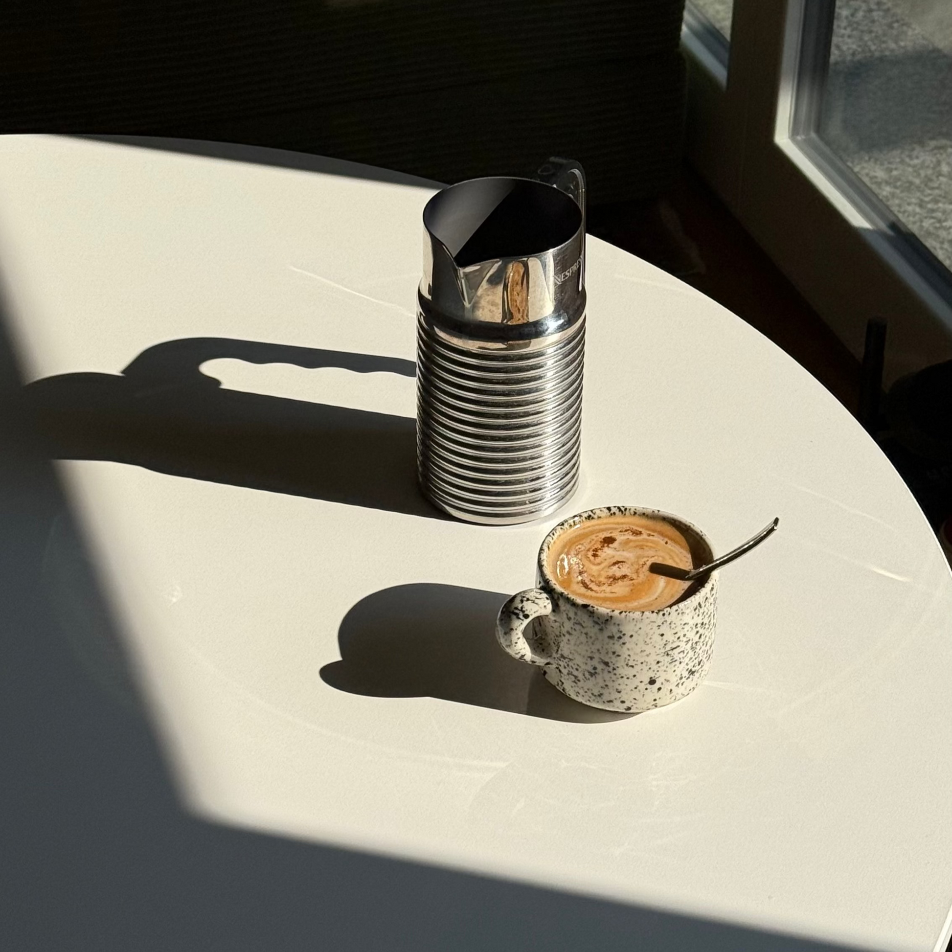 Cup of coffee with a spoon on a white surface, with a metal pitcher in the background. handmade ceramic mug, speckled ceramic