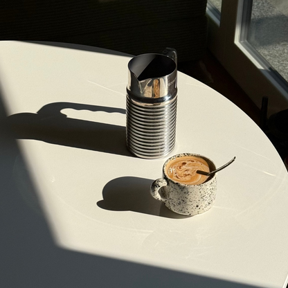 Cup of coffee with a spoon on a white surface, with a metal pitcher in the background. handmade ceramic mug, speckled ceramic