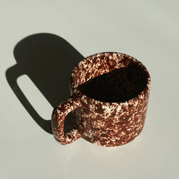 Brown speckled ceramic mug on a textured white surface. Chocolate brown ceramic coffee cup. Handmade ceramic mug.