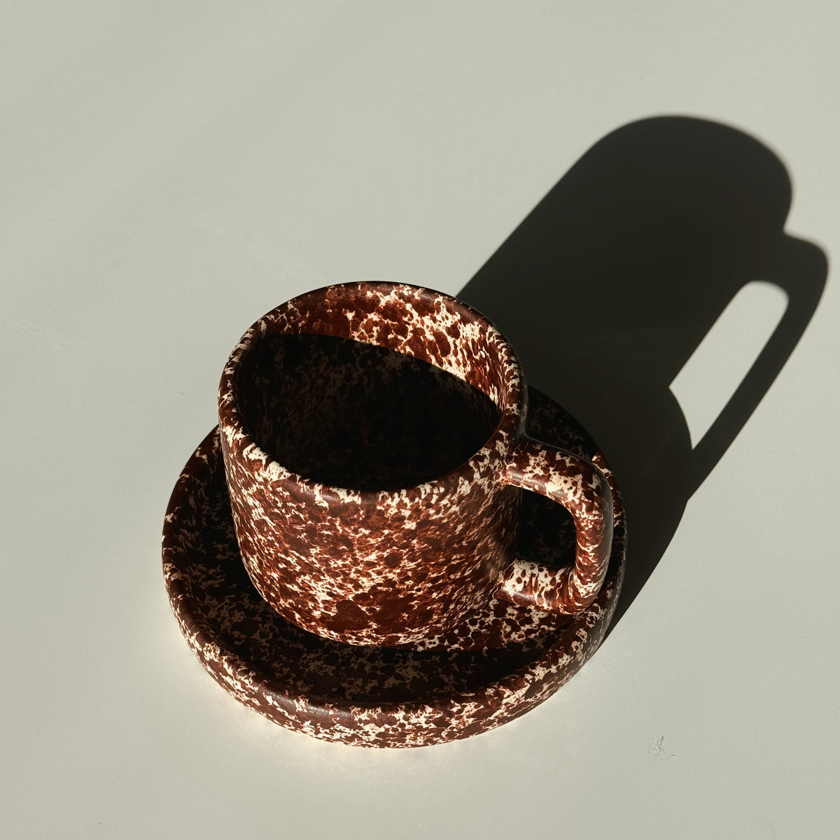 Copper mug and saucer casting a shadow on a light gray background. Handmade ceramic mug set, burgundy, fall colors. Handmade coffee mug.