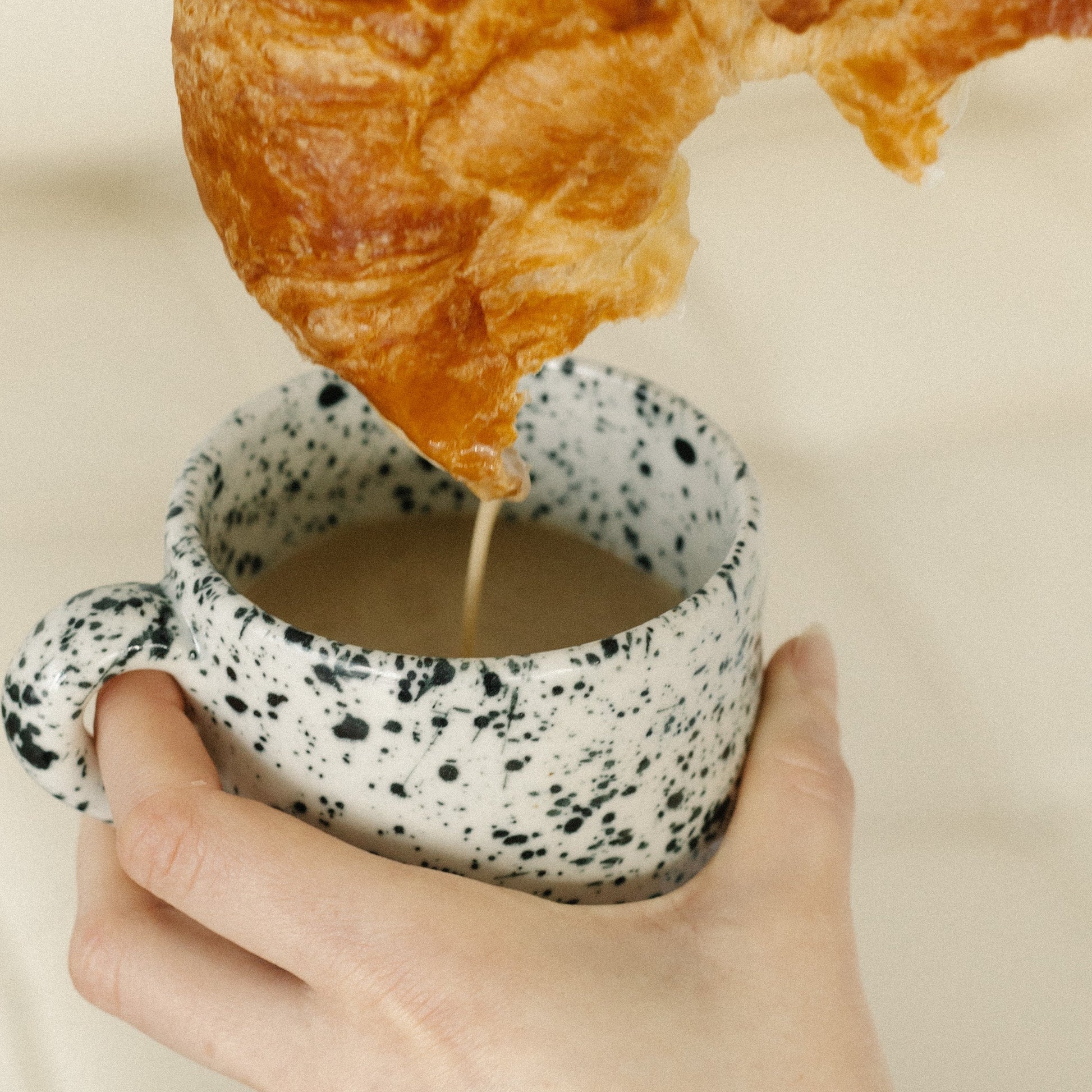 Hand holding a speckled ceramic mug with a croissant in the background